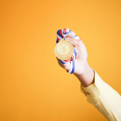 Hand hält eine goldene Medaille mit der Zahl 1 an einem rot-weiß-blauen Band vor orangefarbenem Hintergrund
