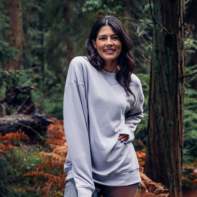 Weibliche Person mit langen dunklen Haaren und grauem Pullover steht in einem Wald mit Farnen und Bäumen im Hintergrund