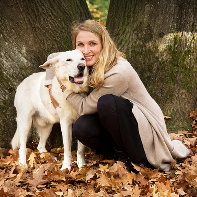 Eine weibliche Person mit langen blonden Haaren hockt im Wald vor einem großen Baum und umarmt einen weißen Hund auf braunem Laubboden
