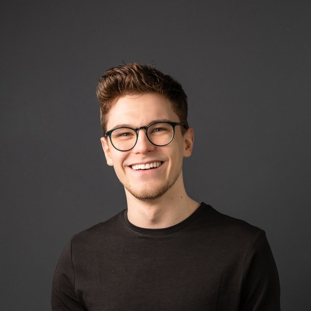 Junger Mann mit braunem Haar trägt ein schlichtes schwarzes T-Shirt und eine Brille vor grauem Hintergrund und lacht in die Kamera