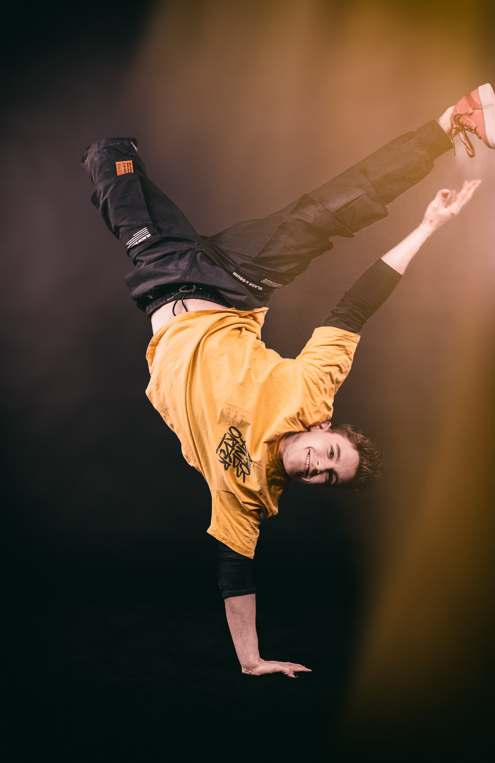 Männliche Person mit kurzen dunkelblonden Haaren macht Handstand auf einer Hand und trägt lockere Hip-Hop-Kleidung