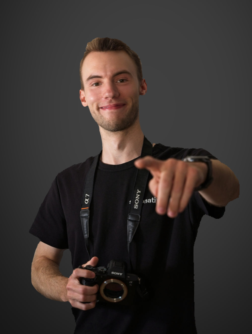 Junger Mann mit kurzem Bart und schwarzem T-Shirt hält eine Kamera und zeigt lächelnd mit dem Finger nach vorne in Richtung Kamera.