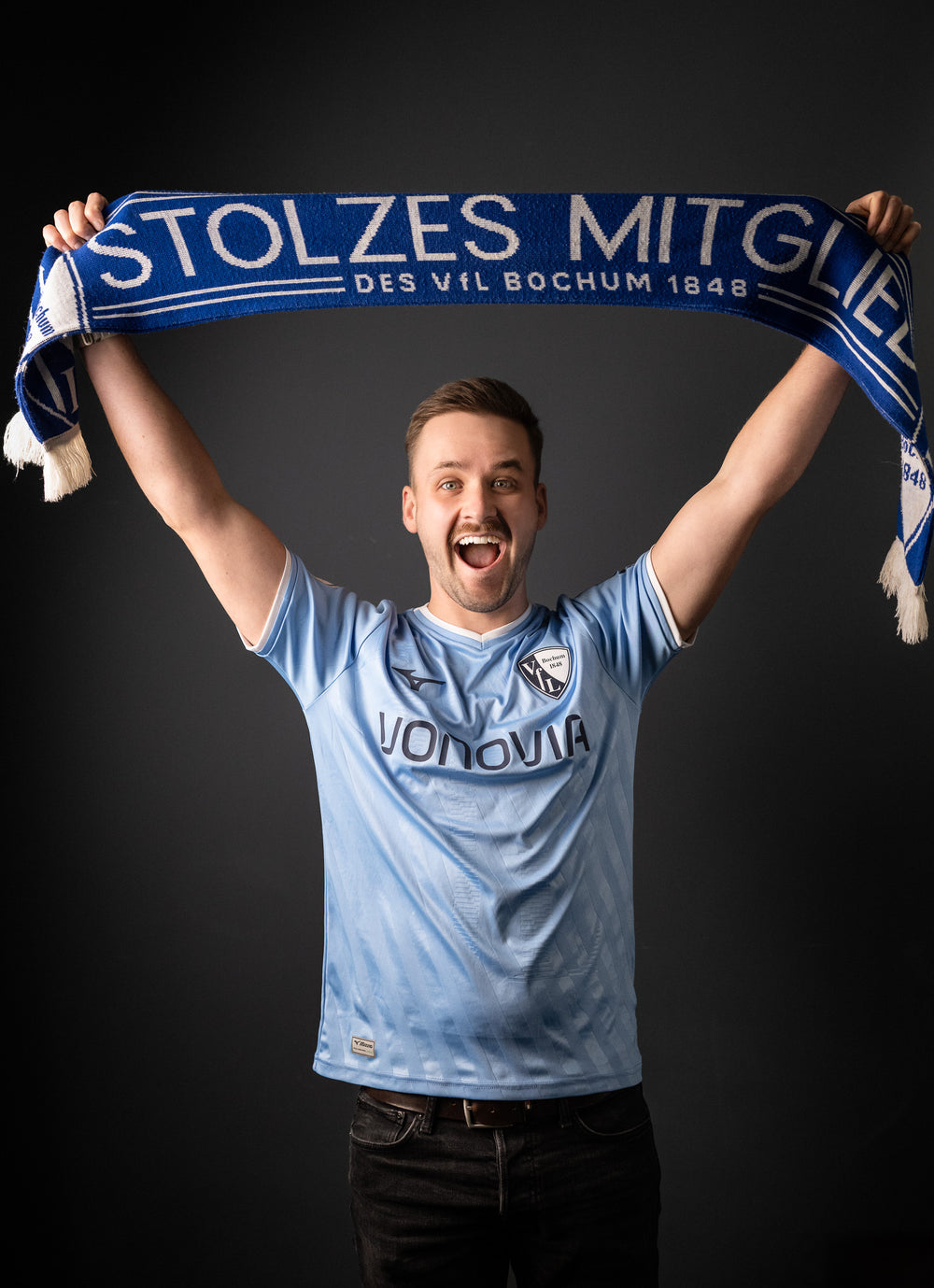 Junger Mann mit hellblauem VfL-Bochum-Trikot hält jubelnd einen blauen Schal mit der Aufschrift 'STOLZES MITGLIED DES VfL BOCHUM 1848' über dem Kopf