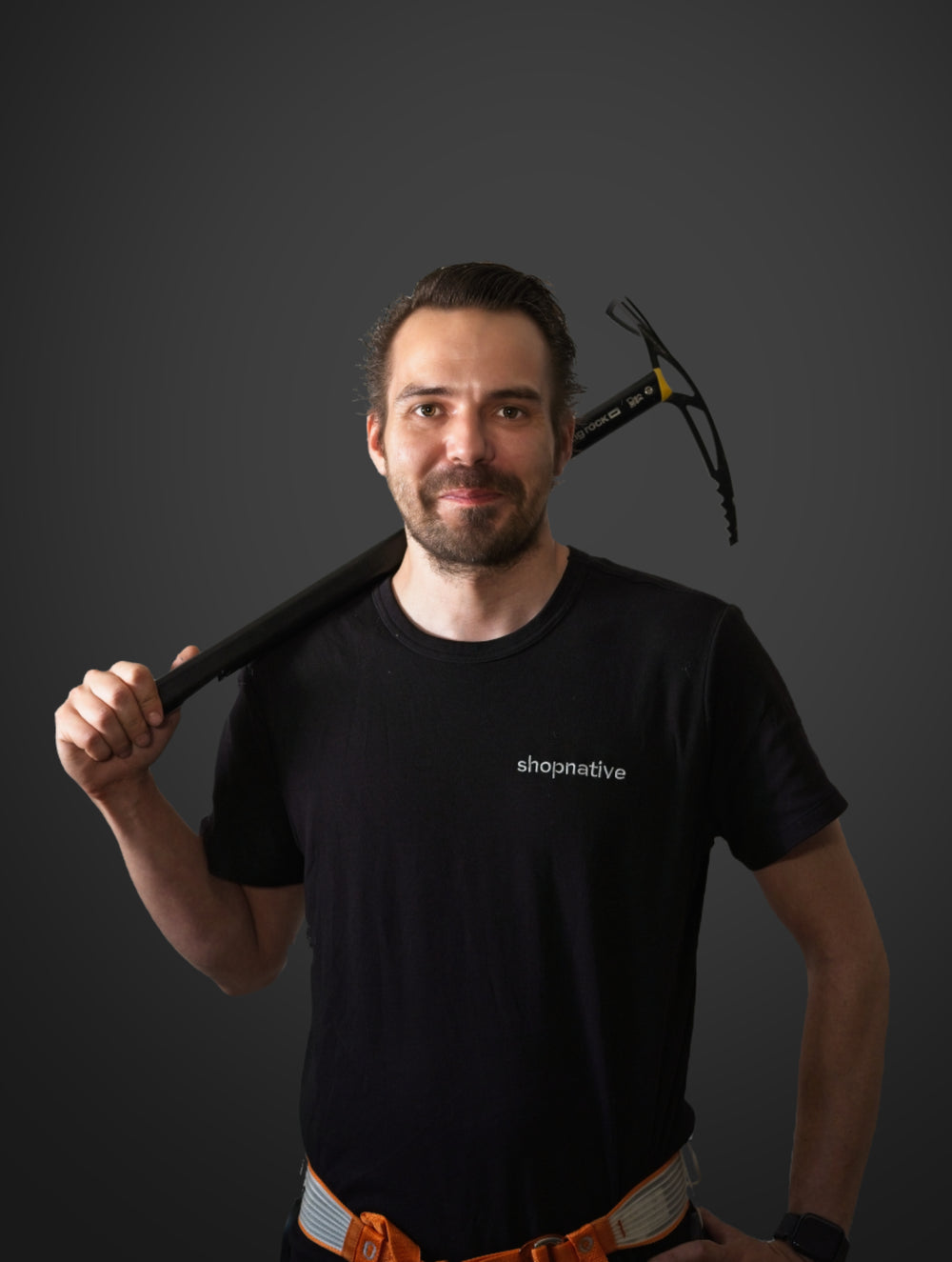 Mann mit kurzem Bart und schwarzem T-Shirt hält eine Eisaxt über der Schulter und trägt Kletterausrüstung. 