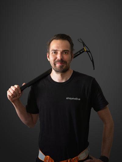 Mann mit kurzem Bart und schwarzem T-Shirt hält eine Eisaxt über der Schulter und trägt Kletterausrüstung. 