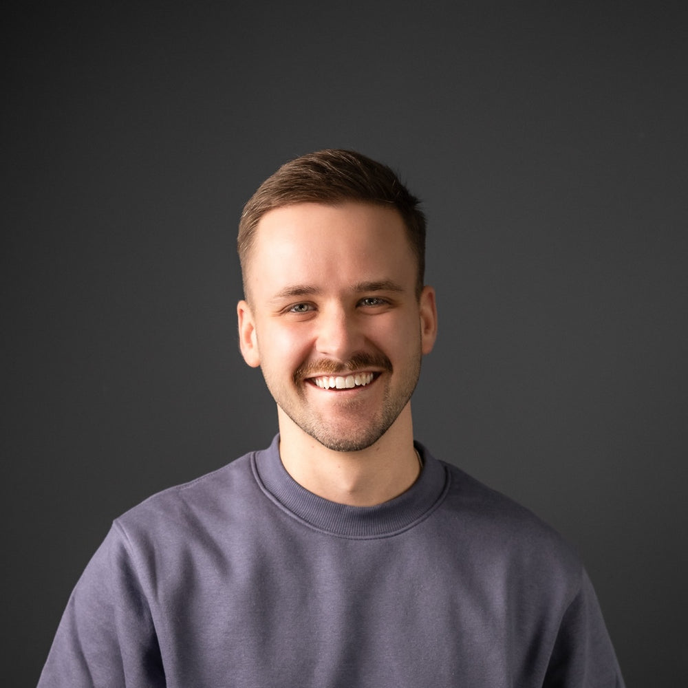Mann mit kurzem Haar trägt ein schlichtes Sweatshirt vor dunklem Hintergrund und grinst breit in die Kamera 