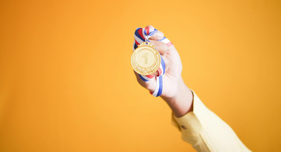 Ein nach oben ausgestreckter Arm hält vor orangenem Hintergrund eine goldene Medaille in die Luft. Das blau-weiß-rot-gestreifte Band der Medaille liegt fest in der Hand. Auf der Medaille ist die Zahl 1 zu sehen. 