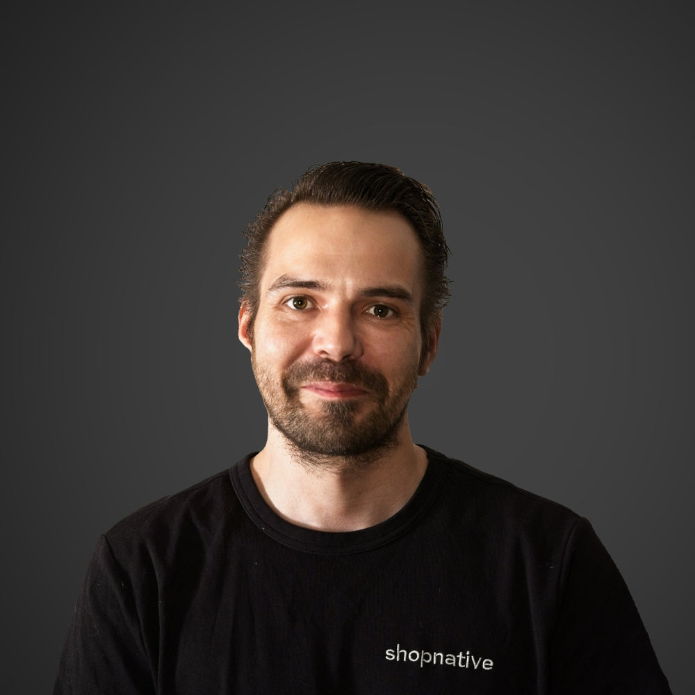 Mann mit kurzen dunklen Haaren und kurzem Bart guckt direkt in die Kamera und trägt ein schwarzes T-Shirt mit der Aufschrift "shopnative".